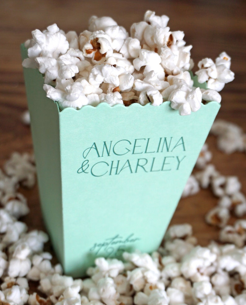 Pu&ograve; includere: Una scatola di popcorn verde chiaro con il testo "Angelina & Charley" stampato sul davanti. La scatola &egrave; piena di popcorn bianchi e circondata da altro popcorn.