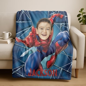 Puede incluir: Una manta azul con un dise&ntilde;o personalizado de Spider-Man. La manta tiene un fondo azul con un gr&aacute;fico de Spider-Man y el nombre "JACKSON" en rojo. La cara de un ni&ntilde;o est&aacute; superpuesta a la cara de Spider-Man.