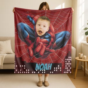 Puede incluir: Una manta roja y azul con tem&aacute;tica de Spider-Man que presenta la cara de un ni&ntilde;o superpuesta en el cuerpo del superh&eacute;roe. La manta tiene un dise&ntilde;o de telara&ntilde;a y el nombre "NOAH" en azul.