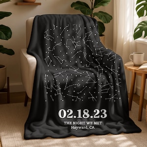 May include: A black throw blanket draped over a chair, featuring a white constellation design. The blanket has the date "02.18.23" and the text "THE NIGHT WE MET Hayward, CA" printed in white. The blanket is made of a soft material.