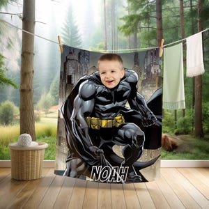 Könnte beinhalten: Ein Batman-Handtuch mit einer Comic-Illustration von Batman mit dem Gesicht eines Kindes. Das Handtuch hat ein Schwarz-Grau-Farbschema mit einem gelben Werkzeuggürtel und dem Namen "NOAH" am unteren Rand. Das Handtuch hängt an einer Wäscheleine.
