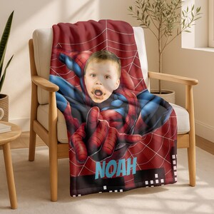 Puede incluir: Una manta roja y azul con tem&aacute;tica de Spider-Man con una imagen personalizada de la cara de un ni&ntilde;o. La manta presenta un dise&ntilde;o de telara&ntilde;a y el nombre "NOAH" en texto azul y blanco.