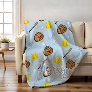 May include: A light blue blanket featuring a softball theme, with illustrations of yellow softballs, brown baseball gloves, and wooden bats. The blanket also has white stars, blue bows, and the name "Karsyn".