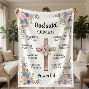 May include: White blanket with floral accents in pink, blue, and peach. The blanket features a wooden cross with floral embellishments and text that reads "God said Olivia is" followed by inspirational words and Bible verses.
