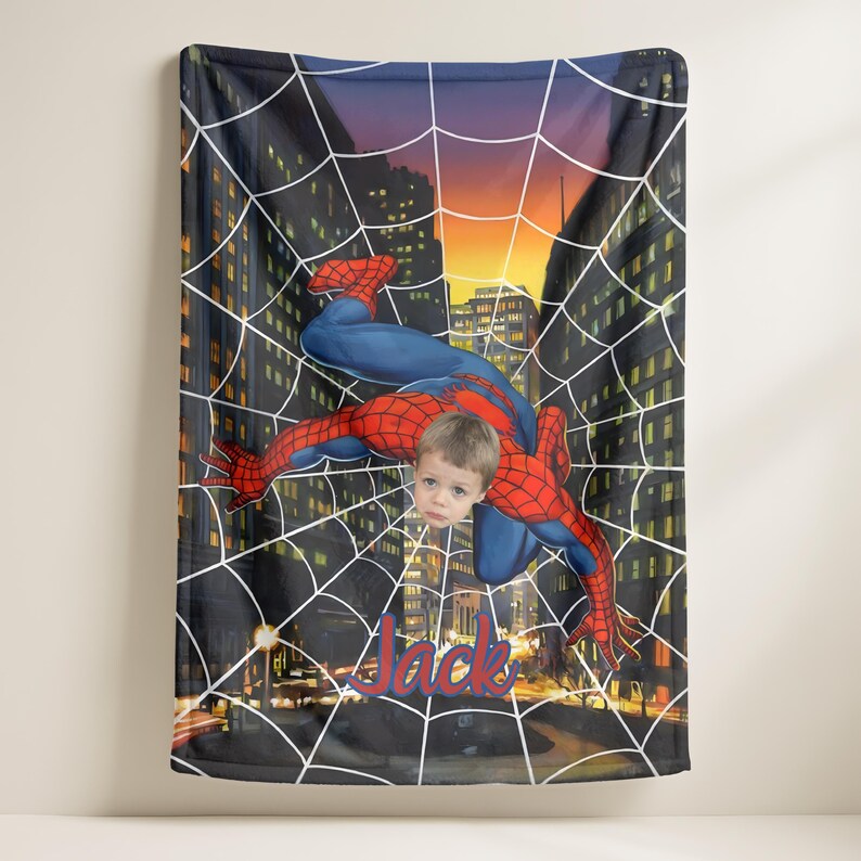 Puede incluir: Una manta personalizada con un dise&ntilde;o de Spiderman con la cara de un ni&ntilde;o superpuesta al personaje. La manta tiene un fondo de paisaje urbano con un dise&ntilde;o de telara&ntilde;a y el nombre "Jack" en escritura roja.