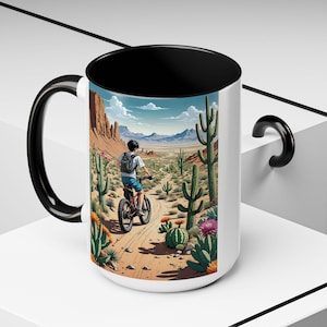 May include: White ceramic mug with a black interior, handle, and rim. The mug features a vibrant illustration of a person riding a bicycle through a desert landscape with cacti, mountains, and a blue sky.