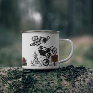 May include: White enamel mug featuring black illustrations of motocross riders performing stunts. The mug has a silver rim and a white handle. The background is blurred, suggesting an outdoor setting.