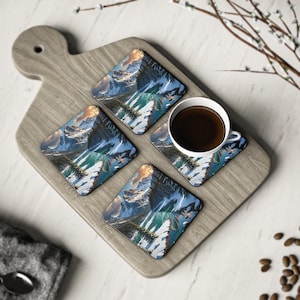 Puede incluir: Cuatro posavasos cuadrados con un diseño de paisaje de montaña, colocados en una tabla de servir de madera. Una taza blanca llena de café oscuro se encuentra sobre uno de los posavasos. Los posavasos presentan una paleta de colores azul, blanco y verde.