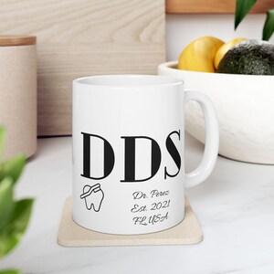May include: White ceramic mug featuring the letters "DDS" in black, a tooth graphic, and text that reads "Dr. Perez Est. 2021 FL USA". The mug is on a beige coaster.