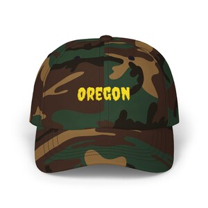 May include: A camouflage baseball cap with the word "OREGON" embroidered in yellow block letters. The cap features a classic six-panel design with a curved brim. The camouflage pattern includes shades of green, brown, and tan.