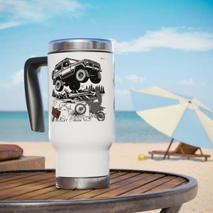 May include: White travel mug with a black handle and a silver lid. The mug features a black and white graphic of an off-road vehicle, a dirt bike, and a sign. The mug is sitting on a wooden table on a beach.