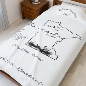 Puede incluir: Manta blanca con un contorno negro del estado de Minnesota, con el texto "Minnesota Est 1858". La manta también incluye las frases "The North Star State" y "Happy 250th, America!"