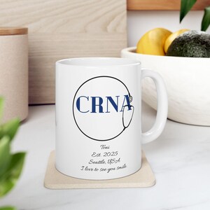 May include: White ceramic mug with a black circular design containing the letters "CRNA" in navy blue, and a stethoscope graphic. Below the circle are the words "Toni Est. 2025 Seattle, USA I love to see you smile."