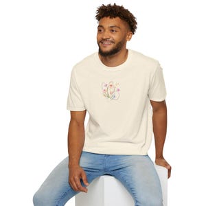 May include: Cream-colored t-shirt with a small floral design in the center. The design features pink, purple, and blue flowers with green stems and leaves. The shirt is paired with blue jeans.