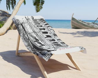 May include: A white beach chair with a black and white illustration of a boat on the seat. The chair is made of light-colored wood and is set on a sandy beach with a boat in the background.