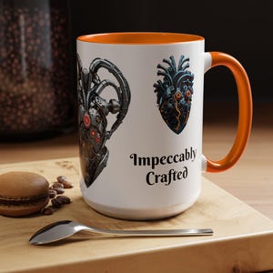 May include: White ceramic mug with an orange interior and handle. The mug features two illustrations of mechanical hearts and the text "Impeccably Crafted" in black. A spoon and a macaron are on a wooden surface.