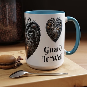 May include: White ceramic mug with a blue interior and handle. The mug features two steampunk-style heart designs with gears and mechanical elements. The words "Guard It Well" are printed below the hearts.