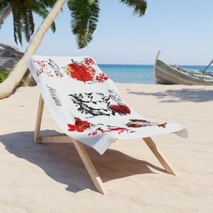 Könnte beinhalten: Ein weißer Strandstuhl mit einem Aufdruck mit roten und schwarzen Blumen- und Koi-Fisch-Illustrationen. Der Stuhl ist aus hellem Holz gefertigt und steht an einem Sandstrand mit einem Boot im Hintergrund.