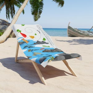 May include: A beach chair with a white fabric seat featuring a river scene with koi fish, leaves, and text. The chair is made of light-colored wood and is set on a sandy beach with a boat in the background.