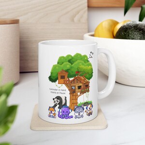 Puede incluir: Taza de cerámica blanca con una colorida ilustración de una casa en el árbol y animales de dibujos animados. El texto "Controller in Hand, World on Pause" está impreso en la taza. La taza está sobre un posavasos beige.