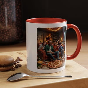 Könnte beinhalten: Weißer Keramikbecher mit roter Innenseite und Henkel. Der Becher zeigt eine farbenfrohe Illustration von Menschen, die einen Horrorfilm ansehen, mit Pizza und Snacks auf einem Tisch. Der Becher steht auf einer Holzoberfläche.