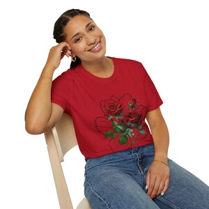 Puede incluir: Una camiseta roja con un gráfico de rosas rojas y hojas verdes. Las rosas están sobre un contorno negro de un lazo. La modelo lleva vaqueros azules y joyas doradas.
