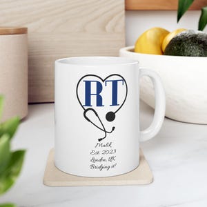 Puede incluir: Taza de cerámica blanca con un diseño de corazón azul con las letras "RT" y un gráfico de estetoscopio. La taza también incluye el nombre "Malik", el año "2023" y la ubicación "London, UK". La frase "Bridging it!" también está impresa en la taza.