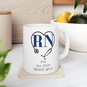 May include: White ceramic mug with a blue heart design containing the letters "RN" and a stethoscope illustration. The mug also features the name "Coni", the year "Est. 2025", and "BMCC, USA" in a script font.