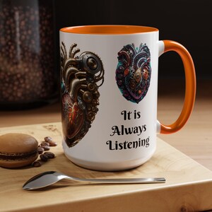 May include: White ceramic mug with an orange interior and handle. The mug features two detailed illustrations of mechanical hearts and the text "It is Always Listening" in black script. The mug is on a wooden surface with a spoon and a macaron.