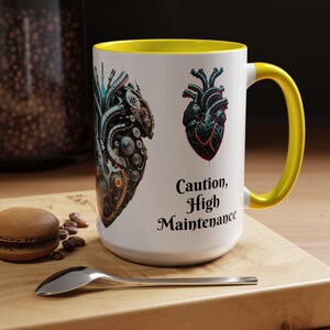 May include: White ceramic mug with a yellow interior and handle. The mug features two detailed illustrations of mechanical hearts and the text "Caution, High Maintenance" in black script. A spoon and a pastry are on a wooden surface.
