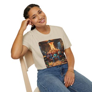 Könnte beinhalten: Cremefarbenes T-Shirt mit einem rechteckigen Grafikmotiv, das eine Wohnzimmerszene zeigt. Die Grafik zeigt Personen um ein Feuer herum, mit Pizza und Getränken. Das Bild deutet auf ein ungezwungenes Treffen oder eine gesellschaftliche Veranstaltung hin.