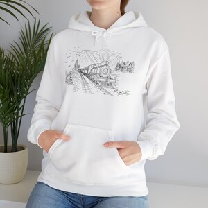 Pode incluir: Moletom branco com capuz com uma ilustração em preto e branco de um trem a vapor viajando por uma paisagem montanhosa. O design inclui um trem sobre trilhos, montanhas, árvores e uma pequena casa. O texto "M. Heritage" também é visível.