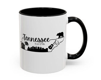 Puede incluir: Taza de cerámica blanca con interior y asa negros. La taza presenta un diseño de silueta negra del estado de Tennessee, que incluye un oso, una guitarra y el texto "Tennessee Est 1796".