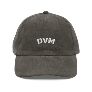 Può includere: Cappellino da baseball in velluto a coste verde oliva con le lettere bianche "DVM" ricamate sul davanti. Il cappellino ha una visiera curva e un bottone rivestito in tessuto. Un accessorio casual.