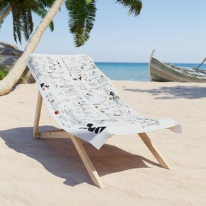 Puede incluir: Una silla de playa blanca con un diseño ilustrado en blanco y negro en la tela. La silla es de madera clara y está colocada en una playa de arena con un barco al fondo. La silla está orientada hacia el espectador.