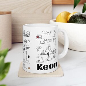 Puede incluir: Taza de cerámica blanca con ilustraciones de arte lineal negro y el nombre "Keon" en letras negras en negrita. La taza presenta varios dibujos de figuras de palo y un signo de interrogación. La taza está sobre un posavasos beige.