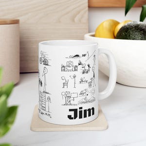 Puede incluir: Taza de cerámica blanca con ilustraciones de figuras de palo negras que representan diversas actividades. La taza tiene el nombre "Jim" impreso en letras negras en negrita. La taza está sobre un posavasos beige.