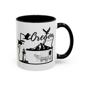 May include: White ceramic mug with a black interior and handle. The mug features a black and white graphic of the state of Oregon, including a whale, eagle, mountain, lighthouse, and wagon. The word "Oregon" is written in script.