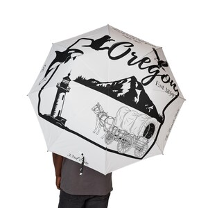 May include: White umbrella featuring black illustrations of Oregon landmarks, including a lighthouse, mountains, and a covered wagon. The word "Oregon" is written in a script font, along with "Est 1859" and "The Beaver State".