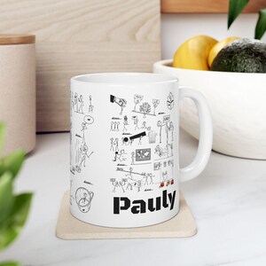 Puede incluir: Taza de cerámica blanca con un diseño de garabatos en blanco y negro. La taza presenta figuras de palitos, bocadillos de diálogo y el nombre "Pauly" en letras negras en negrita. La taza está sobre un posavasos beige.