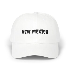 May include: White baseball cap with the text "NEW MEXICO" in black, stylized lettering across the front. The cap has a curved brim and a rounded crown. A casual accessory.