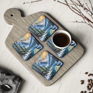 Puede incluir: Cuatro posavasos cuadrados con un diseño de paisaje montañoso, con árboles cubiertos de nieve y un río azul. Una taza de café blanca con líquido oscuro se encuentra sobre un posavasos, colocada sobre una tabla de madera.