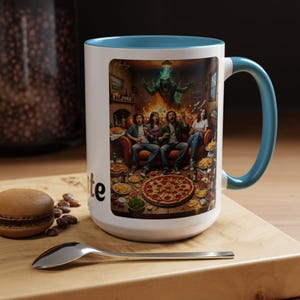Könnte beinhalten: Weißer Keramikbecher mit türkisblauem Innenraum und Henkel. Der Becher zeigt eine farbenfrohe Illustration einer Gruppe von Menschen auf einem Sofa, umgeben von Essen, mit einem Monster im Hintergrund. Das Wort "te" ist auf der Seite sichtbar.