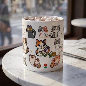 May include: A white ceramic mug filled with a hot beverage topped with marshmallows. The exterior features a repeating pattern of colorful pixel art cats in various poses, a small green turtle, a red flower, and the word Hello in small text.