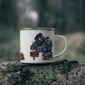 May include: White enamel mug featuring a black silhouette of a motocross rider jumping over a mountain, with the words "Day Off" on a wooden sign. Additional graphics include a tent and a 4x4 vehicle.