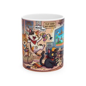 May include: A white ceramic mug featuring a colorful cartoon illustration of three cats playing video games. The design includes text that reads Feline Velocity, Mochi Yaw, Play Hard! Nap Harder!, Vintage Play Unit, and Cat Adventure on a screen.