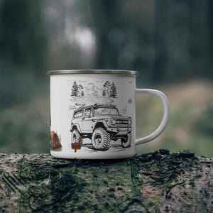 May include: White enamel camping mug with a black illustration of a 4x4 vehicle, trees, mountains, and a tent. The mug has a silver rim and a white handle. The design evokes a sense of adventure and outdoor activities.