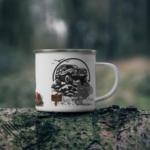 May include: White enamel camping mug featuring a black and white illustration of an off-road vehicle on a rocky terrain, with a motorcycle and text. The mug has a silver rim and a white handle. The background is blurred.