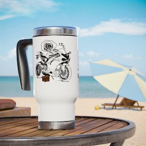May include: White travel mug with a black handle and a silver lid. The mug features a black and white illustration of a skeleton riding a motorcycle. The background shows a beach scene with a blue sky and ocean.