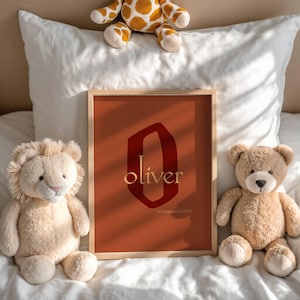 Op de afbeelding: Een ingelijste print met de naam "Oliver" in gouden letters op een roestkleurige achtergrond. De print toont een grote, gestileerde "O" in een donkerdere rode tint. De datum "24 september 2024" staat eronder. Pluche speelgoed omringt de print.